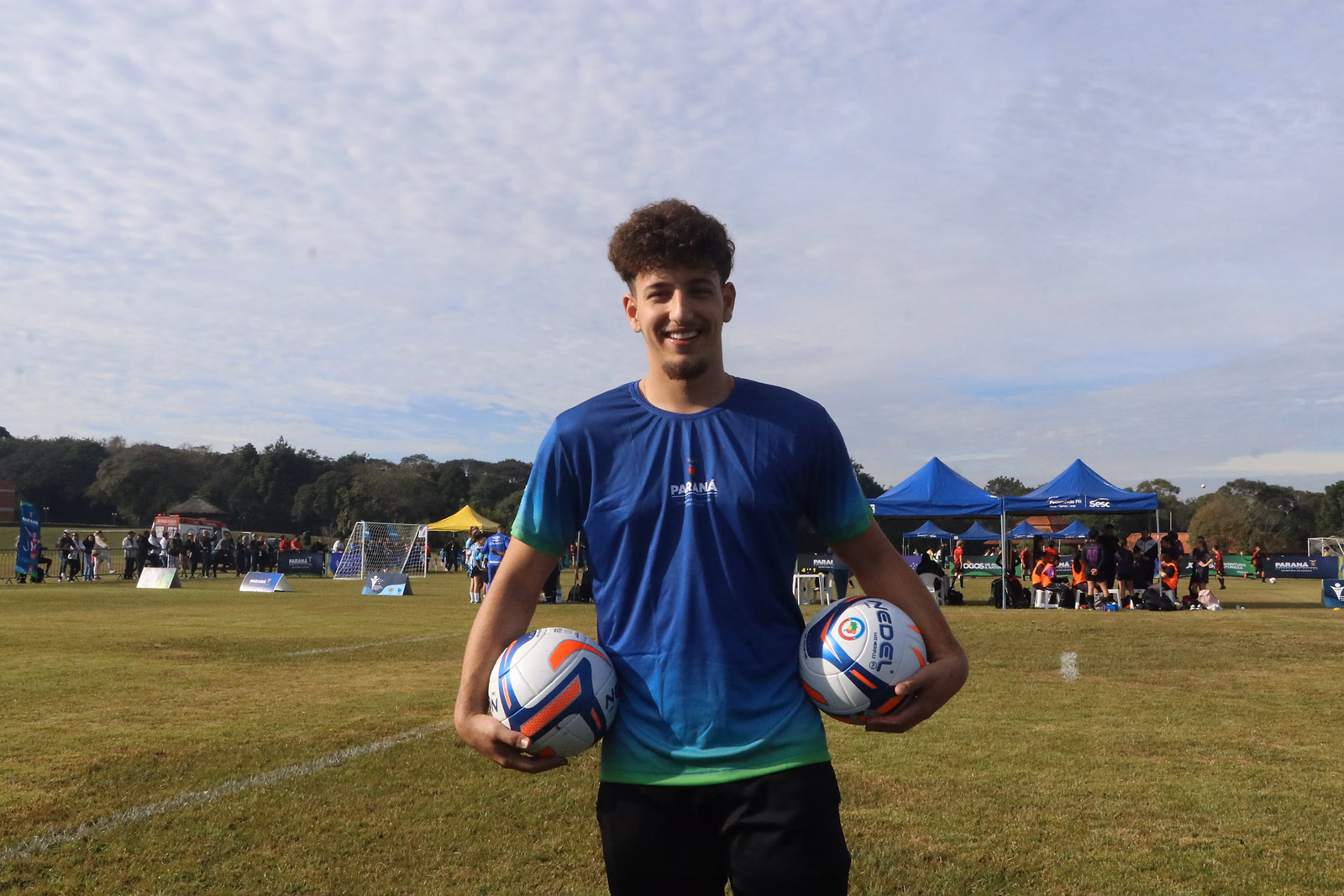Jovem da organização sorri no gramado com as duas bolas do torneio nas mãos, os jogos ao fundo.