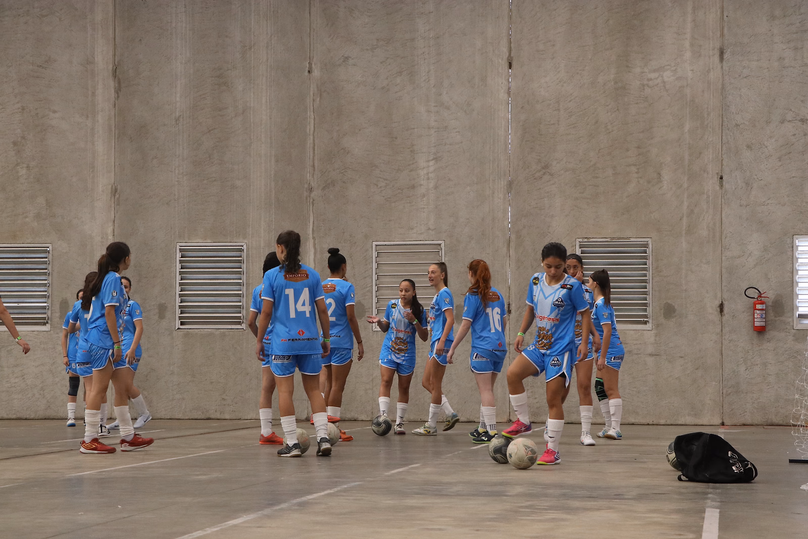 Adolescentes do time feminino aquecem na quadra coberta antes da partida, com as bolas no chão.